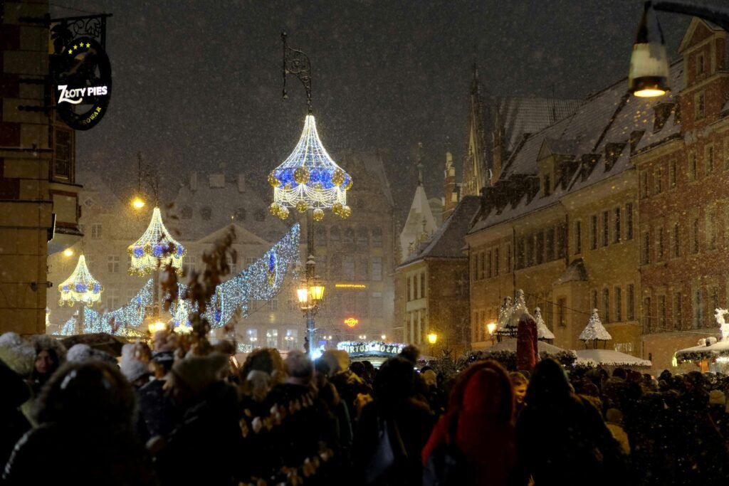 Crowded European city square illuminated with festive holiday lights during a snowy winter night.